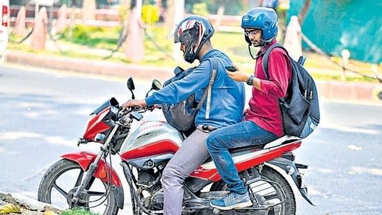 Two men wearing helmets and backpacks sitting on a red motorcycle, with the pillion rider using a smartphone.