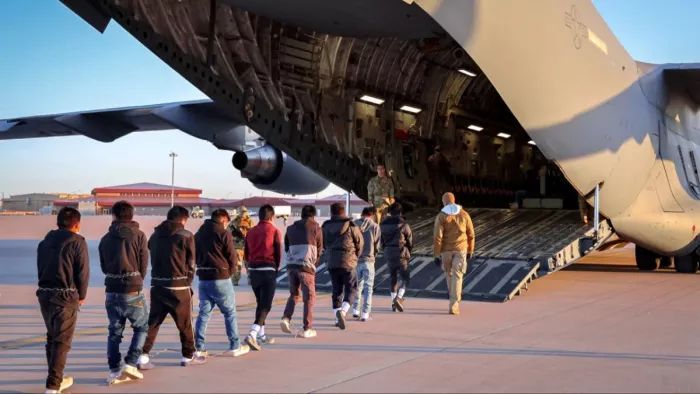 A line of deportees walking up to a large military aircraft ramp, shadows stretching out on the tarmac.