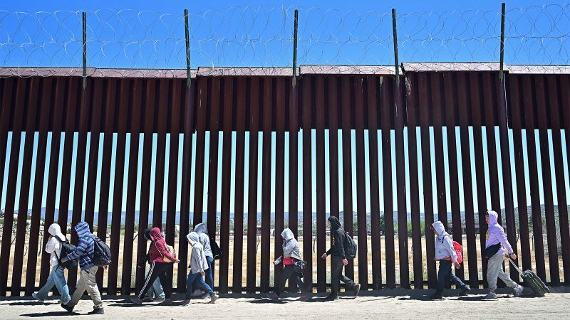 A line of migrants, backpacks on, walk along a border fence under barbed wire in the desert sunlight.