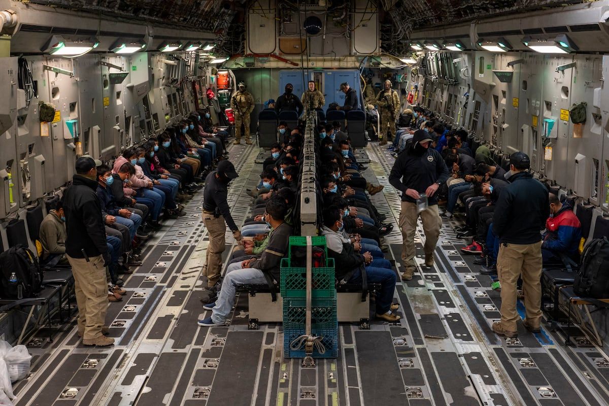 Inside a military cargo plane, rows of deportees seated with handcuffs and escorts around them.